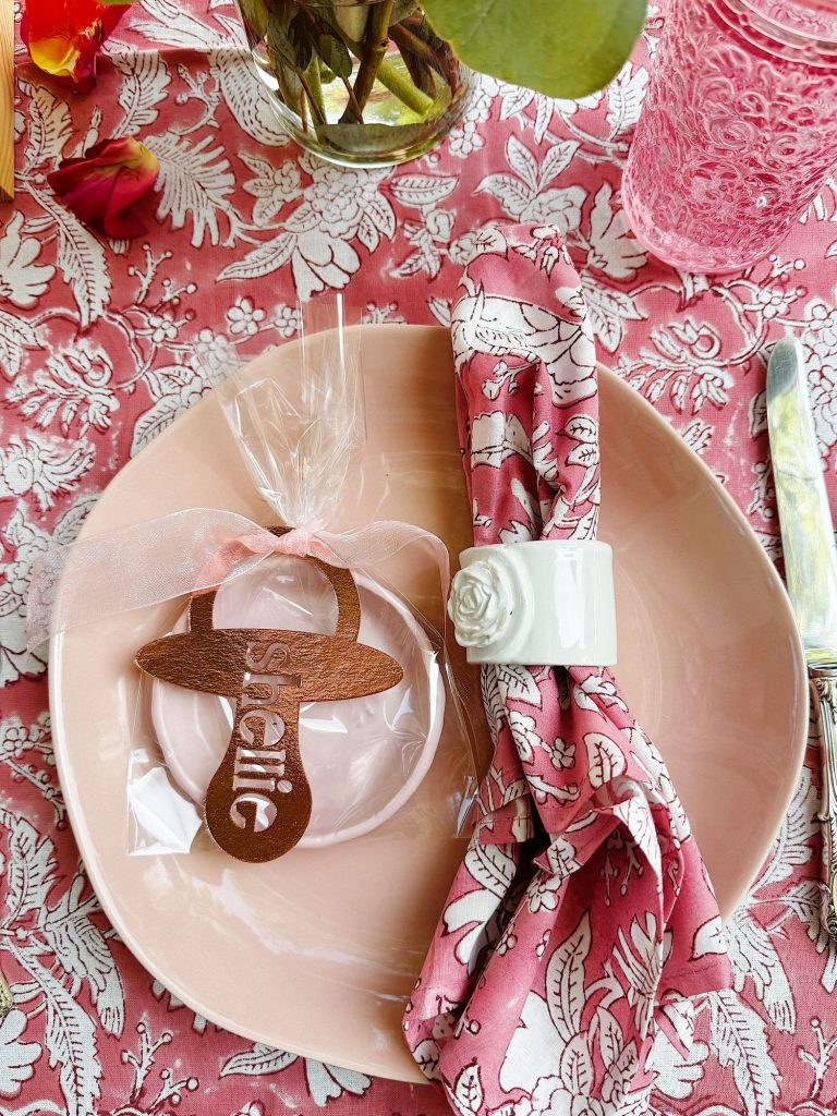 A pink plate with a floral napkin held by a white flower napkin ring, a wooden pacifier-shaped cookie labeled “SAVING,” and decorative glassware on a pink patterned tablecloth.