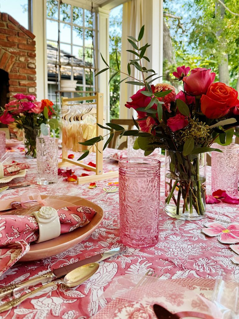 A table set for a meal with pink floral tablecloth, pink glassware, floral centerpieces, and cloth napkins, near large windows with a view of greenery outside.