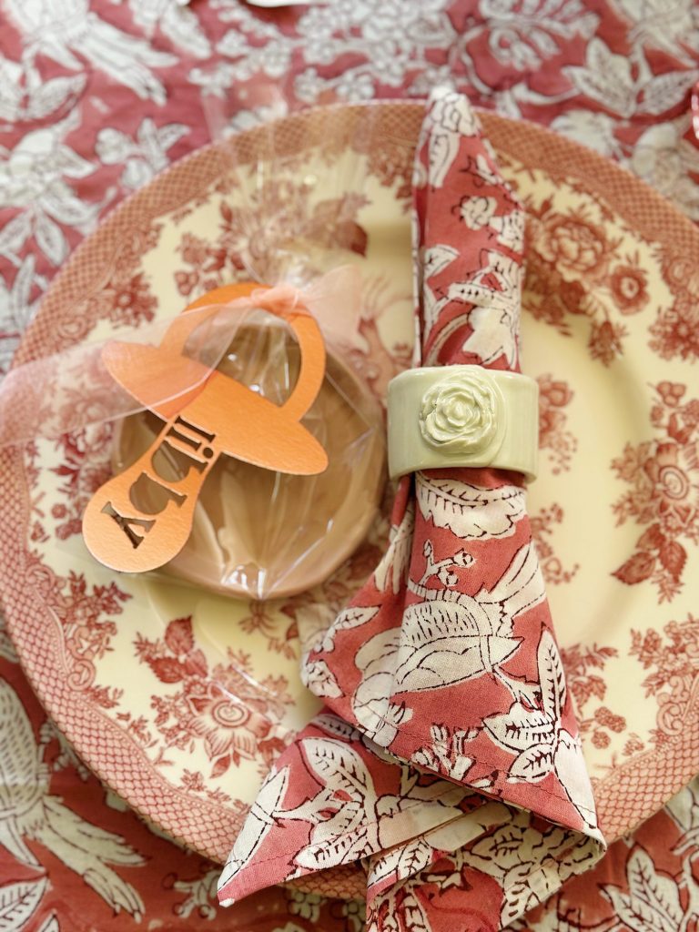 A decorative place setting with a pink and white patterned plate, matching napkin in a white ring, and a pacifier-shaped "baby" tag attached to a wrapped cookie.