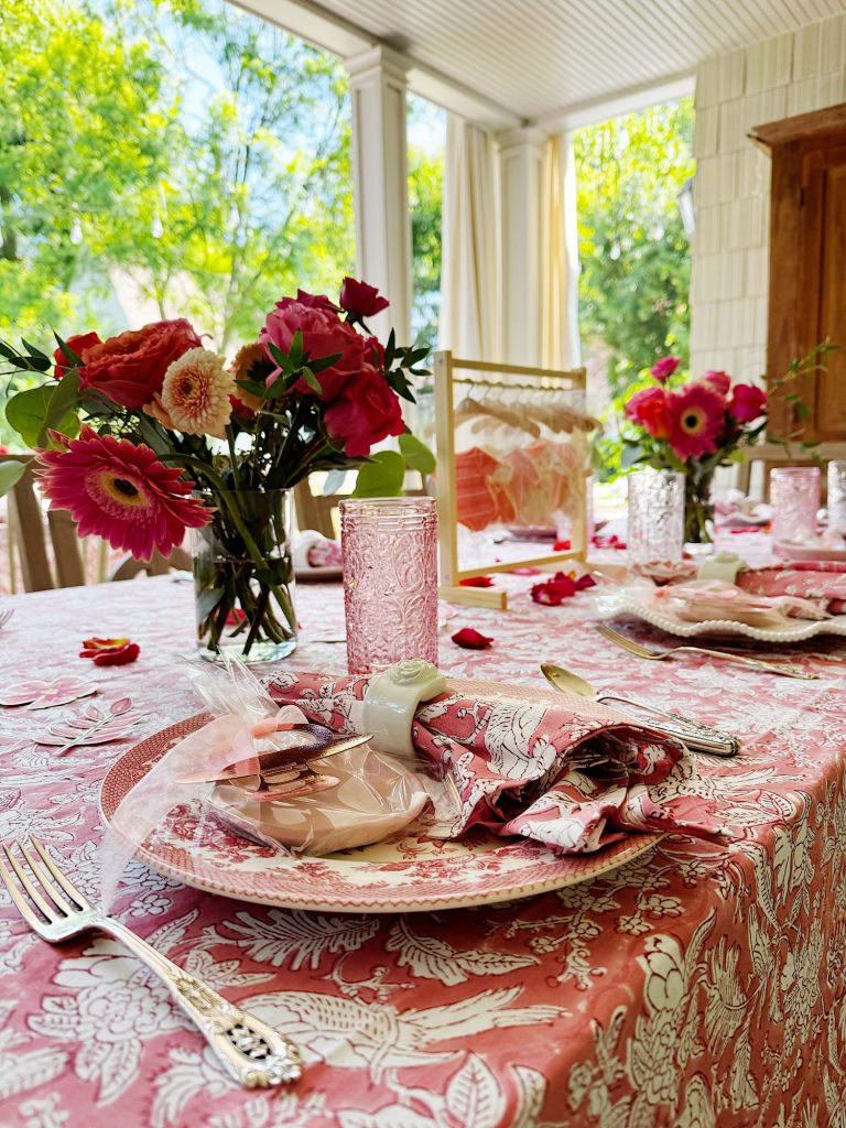 A table set for a meal with pink floral tablecloth, matching plates and napkins, glassware, and vases of red and pink flowers on a covered patio.