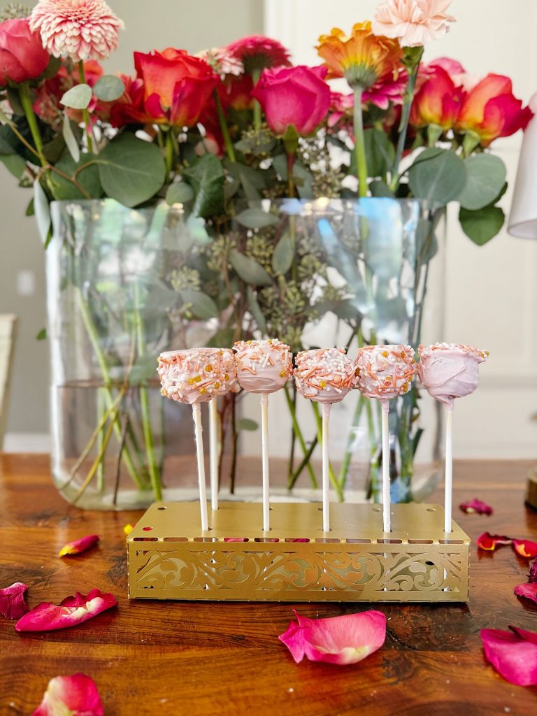 Six pink cake pops with sprinkles on sticks are displayed in a decorative holder on a wooden table, with vases of colorful flowers and scattered petals in the background.