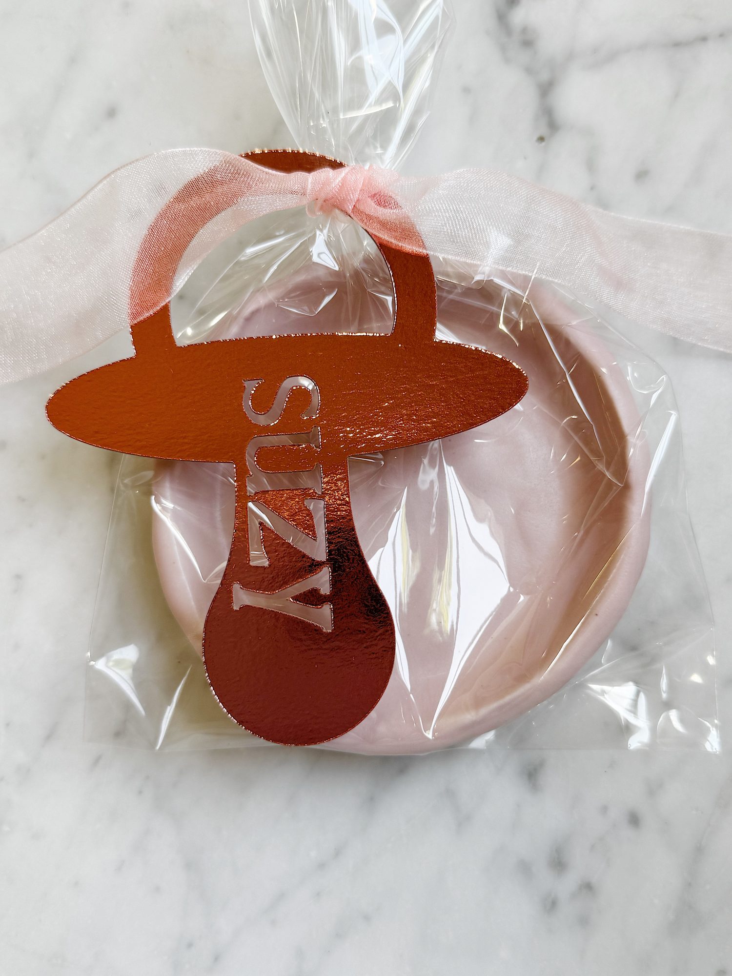 A pink cookie in a clear plastic bag with a metallic pink pacifier-shaped tag labeled &ldquo;Suzy,&rdquo; placed on a marble surface.