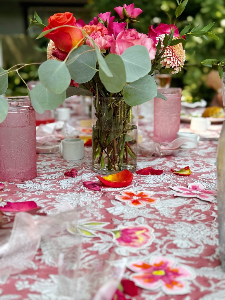 A vase of pink and orange flowers sits on a patterned tablecloth, surrounded by glasses of pink drink, peach slices, and scattered flower petals.
