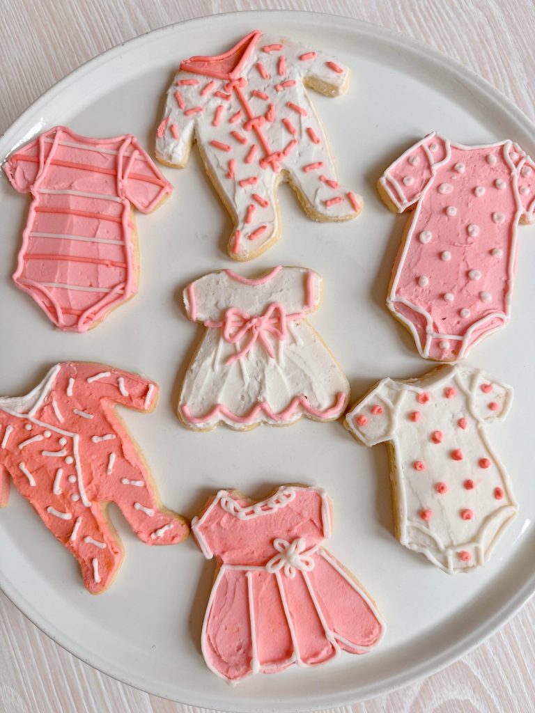 A plate of sugar cookies decorated with pink and white icing in the shapes of baby clothes and dresses.