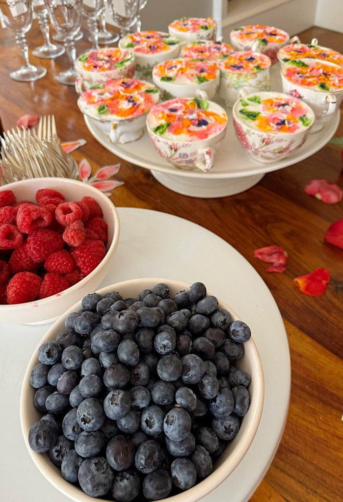 Bowls of blueberries and raspberries sit on a white tray in front of a tiered stand holding decorated teacups filled with dessert, with forks and scattered flower petals nearby.