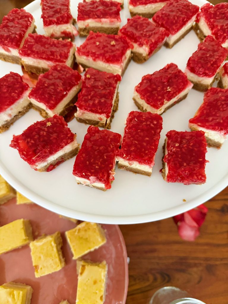 Rectangular raspberry cheesecake bars on a white plate, with yellow dessert bars on a pink plate below, set on a wooden table.