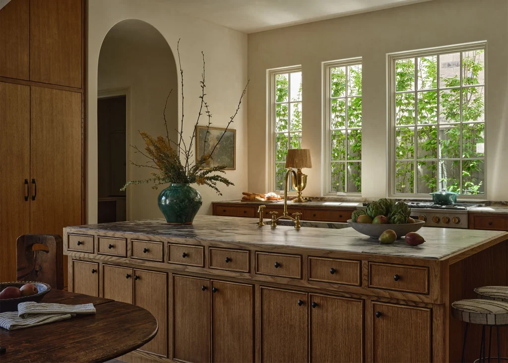 A kitchen with wooden cabinets, marble island countertop, a green vase with branches, a fruit bowl, and large windows letting in natural light.