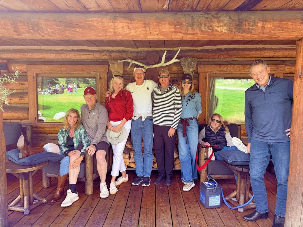 Eight adults pose together on the wooden porch of a log cabin beneath mounted antlers. Some are sitting, others standing, all in casual attire—enjoying each other’s company now that fall is here.