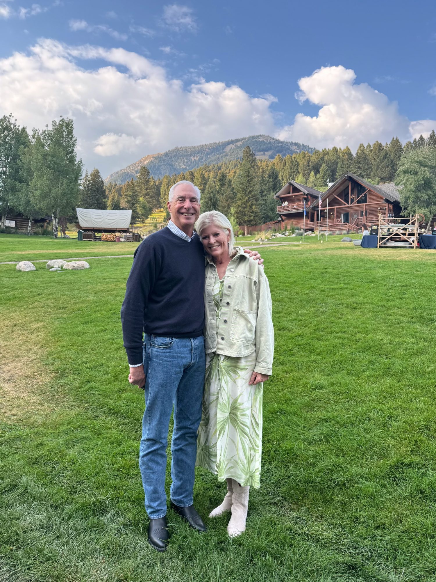 A man and woman stand together on a grassy lawn, smiling, with cabins, trees, and mountains in the background under a partly cloudy sky.