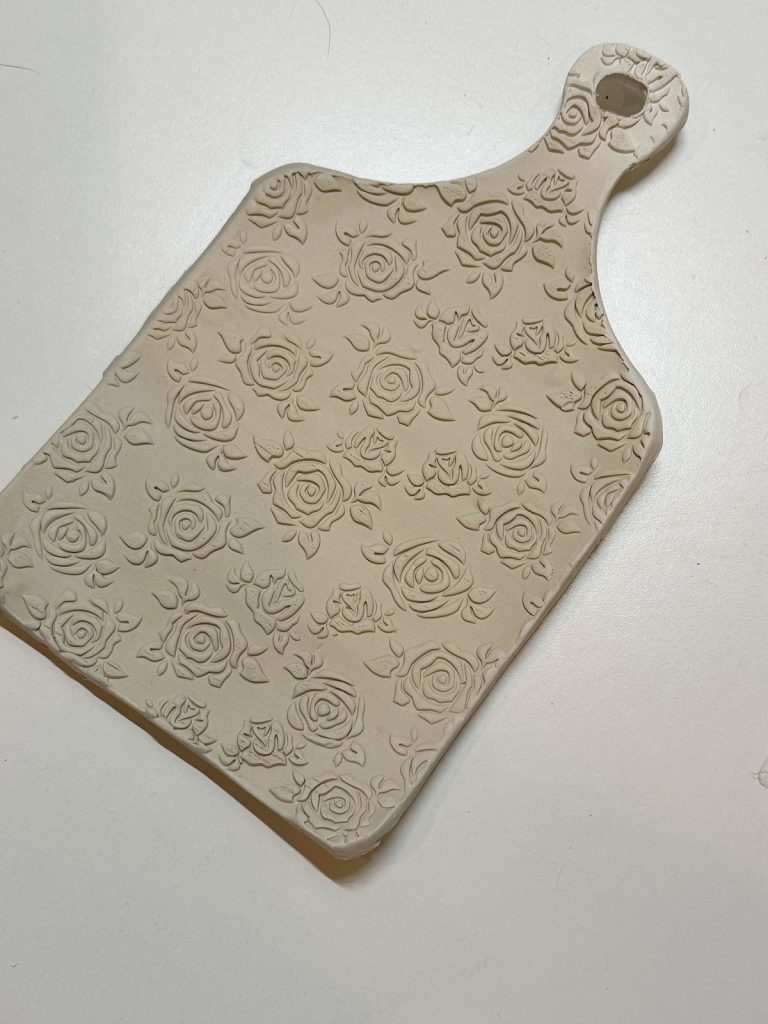 A beige ceramic cutting board with a handle, featuring an embossed rose pattern across its surface, placed on a white background.