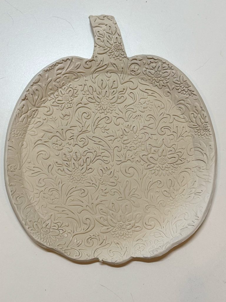 A flat, pumpkin-shaped clay tray with an intricate floral and leaf pattern embossed on its surface, resting on a white background.