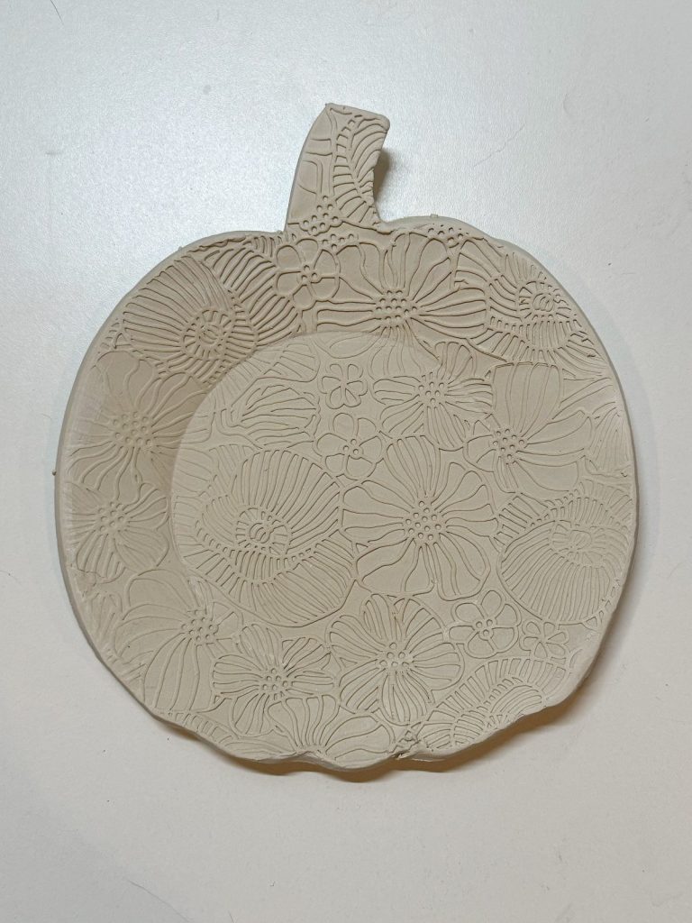 A flat, pumpkin-shaped clay piece with a raised floral pattern covering the surface, placed on a white background.