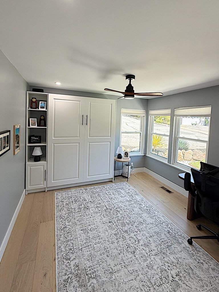 Bright home office with a large window, light wood floor, gray walls, white Murphy bed, shelves, rug, black desk and chair, and ceiling fan.