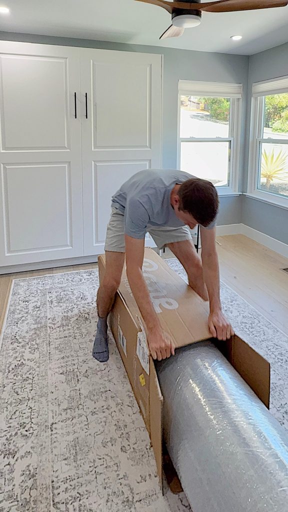 A person opens a large cardboard box containing a rolled-up mattress in a bright, modern room.
