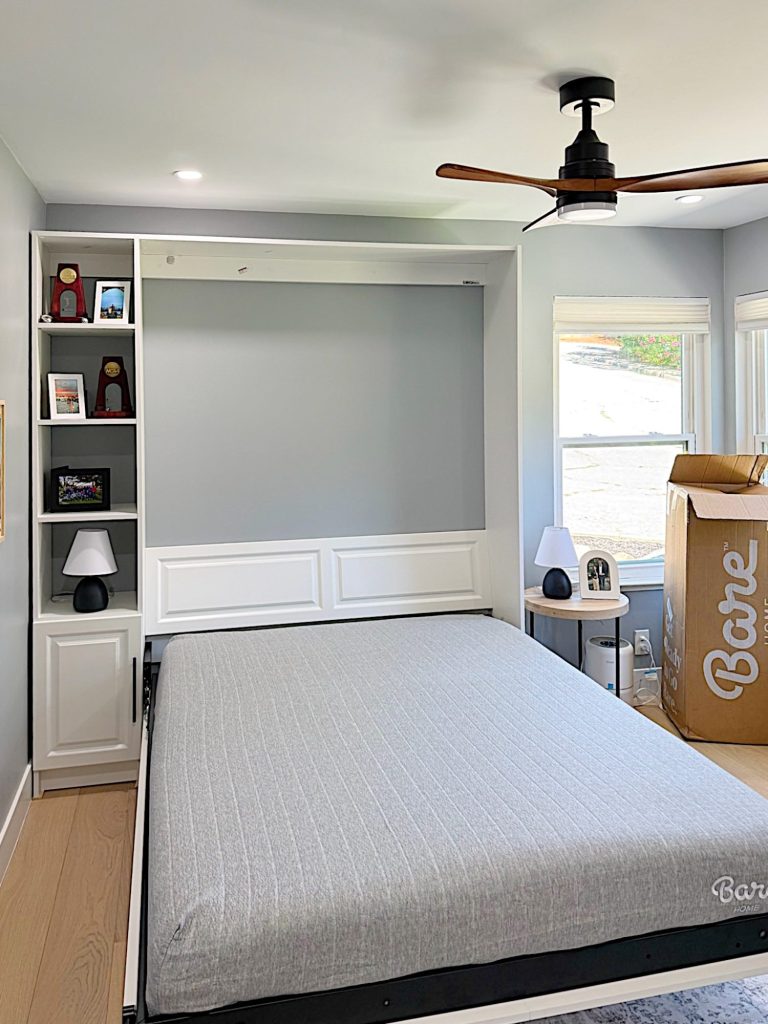 A neatly made Murphy bed is lowered in a modern bedroom with built-in shelves, two lamps, a ceiling fan, and a large cardboard box labeled "Barle" in the corner.