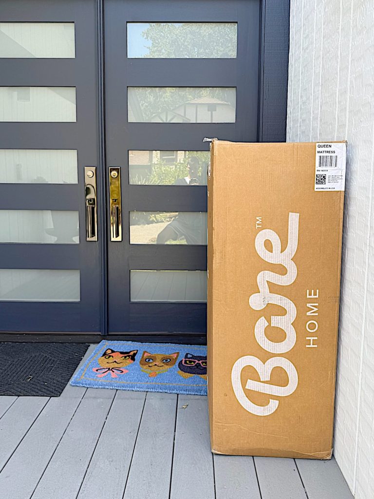 A large Bare Home box is placed upright on a porch near a modern front door, next to a blue doormat with cat faces.