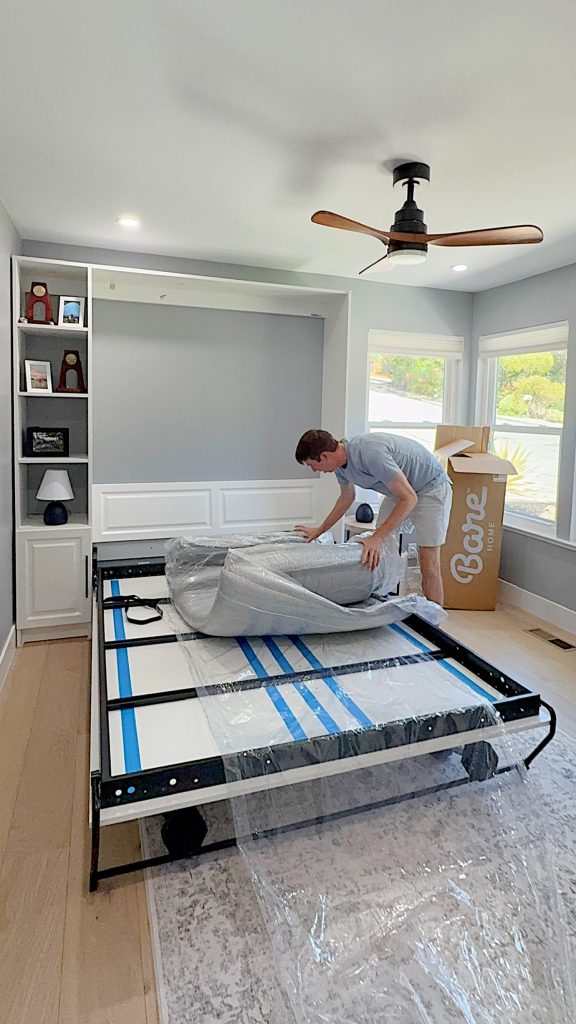 A person unpacks a rolled mattress onto a metal bed frame in a modern bedroom with built-in shelves and a ceiling fan.