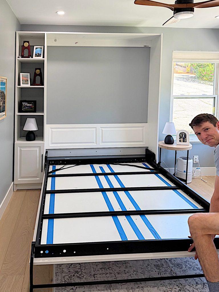 A man assembles a white murphy bed frame in a modern room with built-in shelves, a nightstand, and a window showing a view outside.