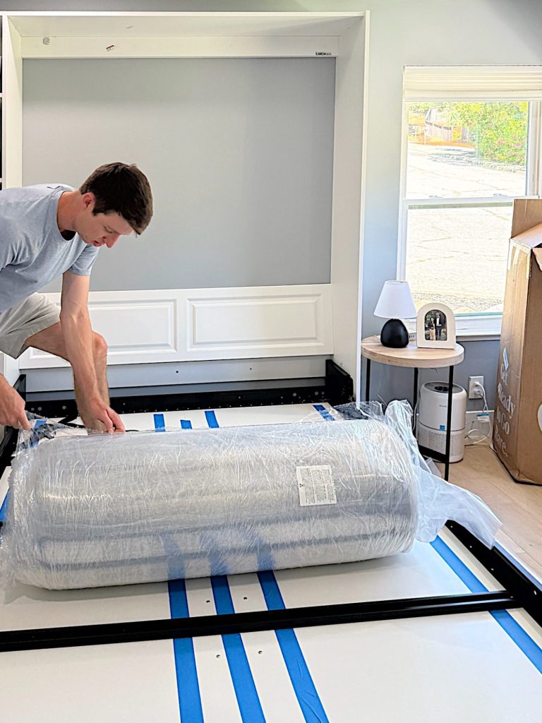 A person cuts open the plastic wrapping of a rolled-up mattress on a partially assembled bed frame in a bright, tidy bedroom.