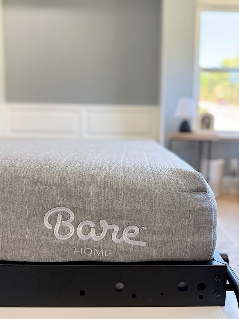 A close-up of a gray mattress with "Bare Home" embroidered on the side, placed on a black metal bed frame in a bright, modern bedroom.