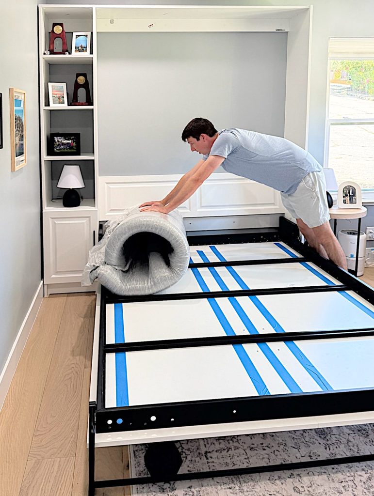 A man in a light shirt and shorts unrolls a mattress onto the frame of a Murphy bed in a bright room with built-in shelves and light wood flooring.