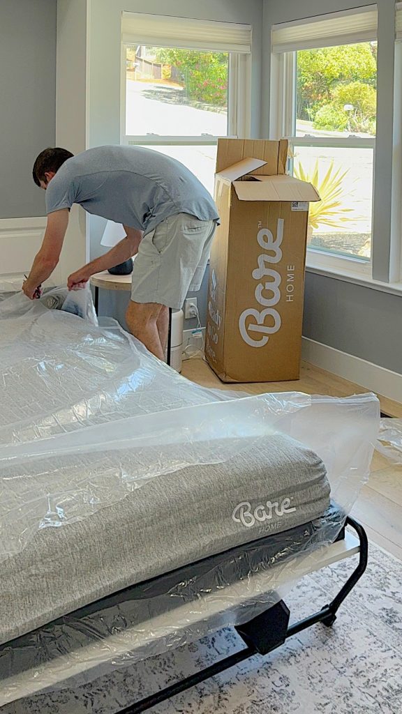 A person unpacks and sets up a Bare Home mattress in a bright bedroom with large windows and a cardboard box nearby.