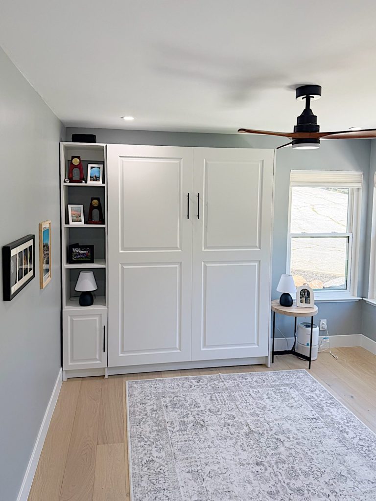 A tidy room with light wood floors, a white cabinet with shelves, a rug, a ceiling fan, and a window letting in natural light.