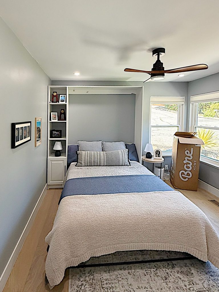 A neatly made bed with striped pillows is centered in a small, modern bedroom with light gray walls, built-in shelves, a ceiling fan, and a large cardboard box by the window.