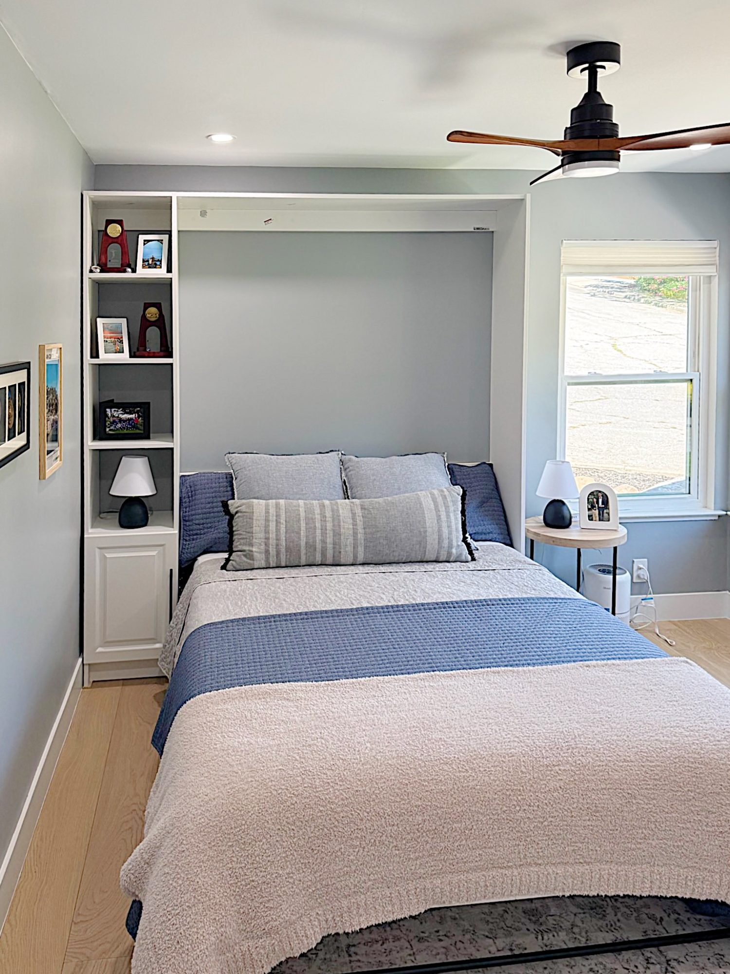 A neatly made bed with blue and gray bedding is centered between built-in shelves and a side table, in a bright bedroom with light walls and a ceiling fan.