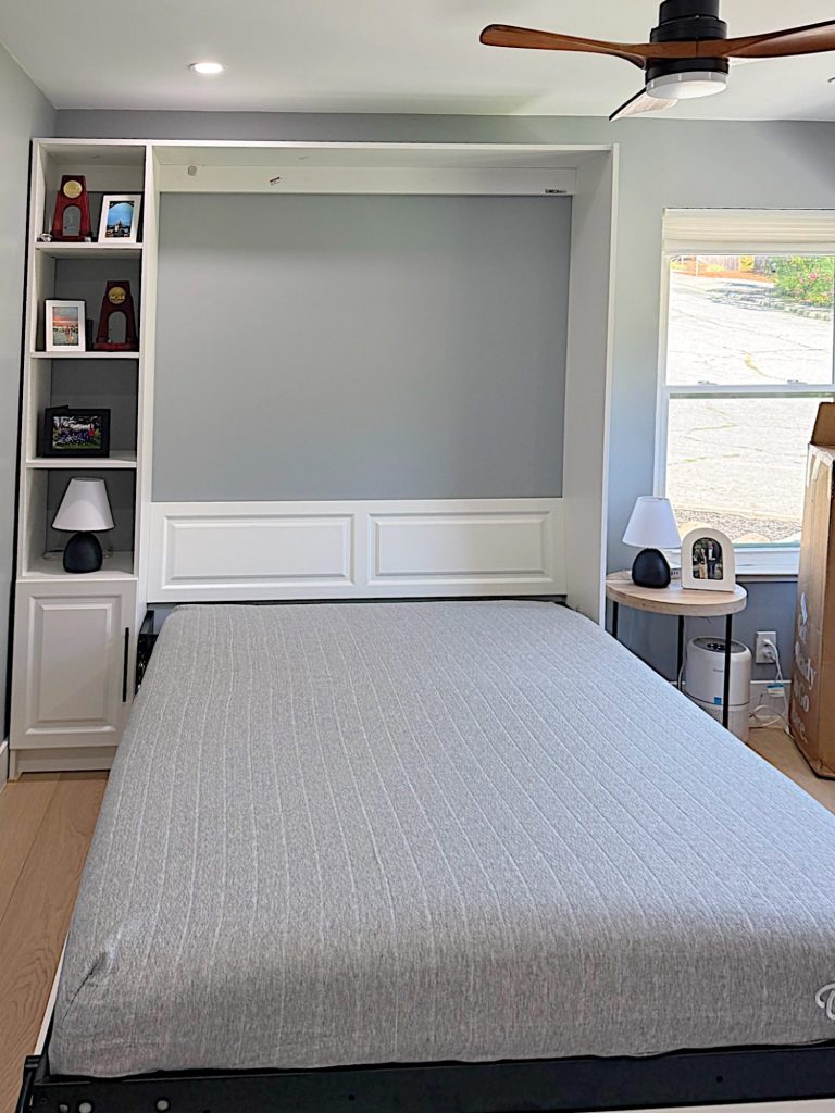 A modern room with a Murphy bed pulled down, gray bedding, built-in shelves with decor and lamps, a ceiling fan, and a window letting in natural light.