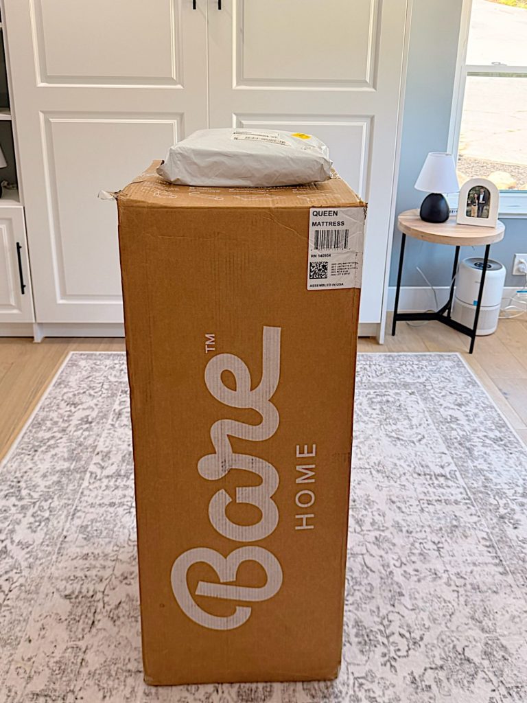 A Bare Home branded brown box labeled "Queen Mattress" stands upright on a rug, with a white package placed on top, in a bright, modern room.