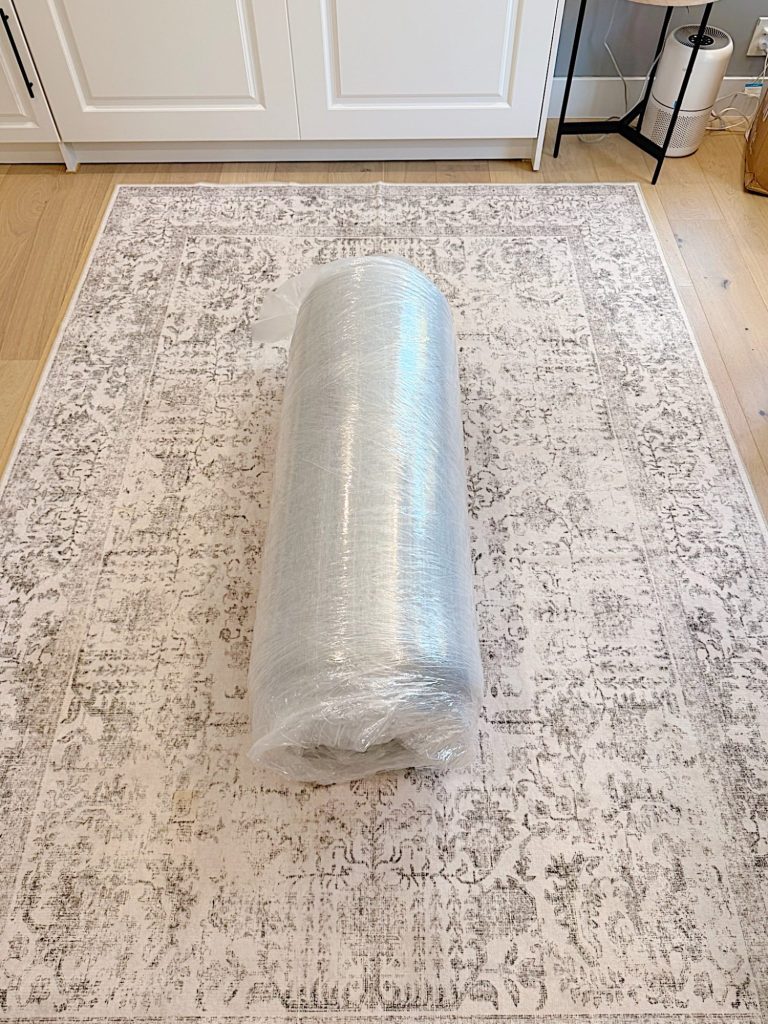 A large rolled-up mattress wrapped in plastic sits on a patterned area rug in a room with light wood floors and white cabinets.
