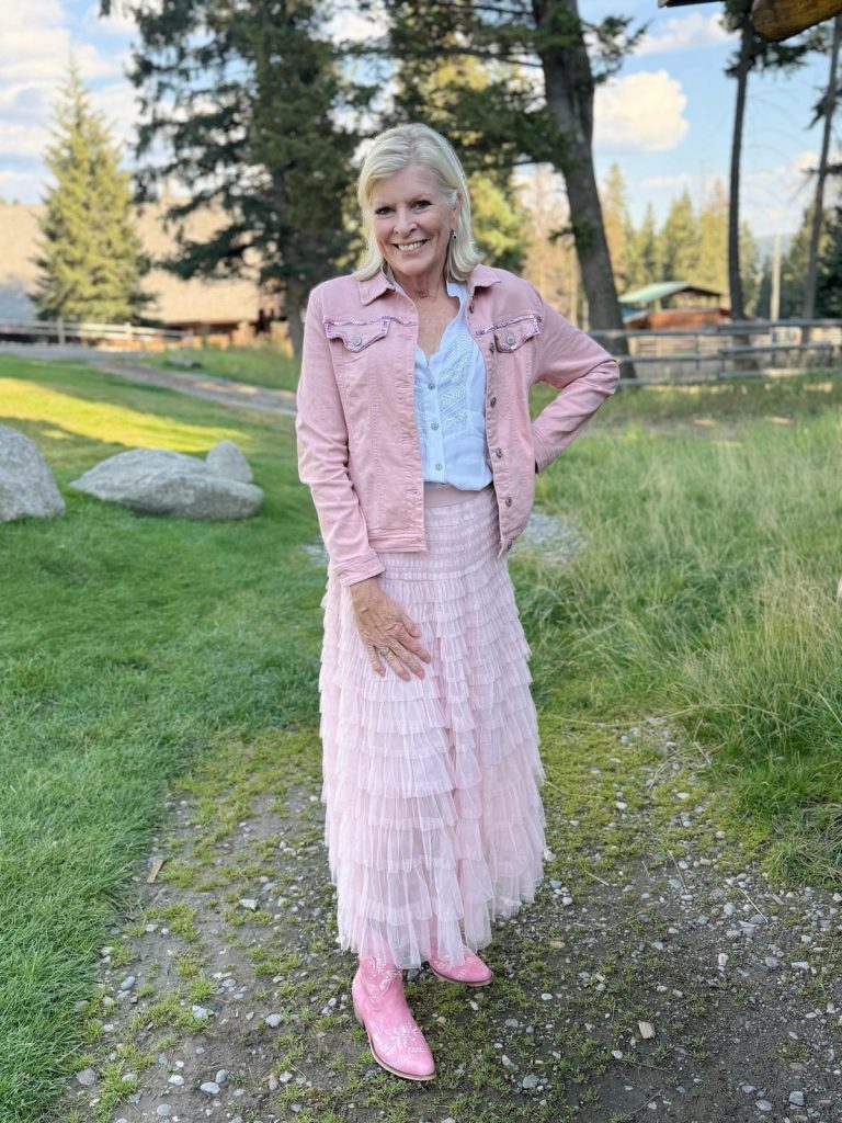 A woman stands outdoors on a gravel path, wearing a light pink jacket, white top, layered pink skirt, and pink boots. Fall is here, with trees, grass, and a wooden fence creating a cozy seasonal backdrop.