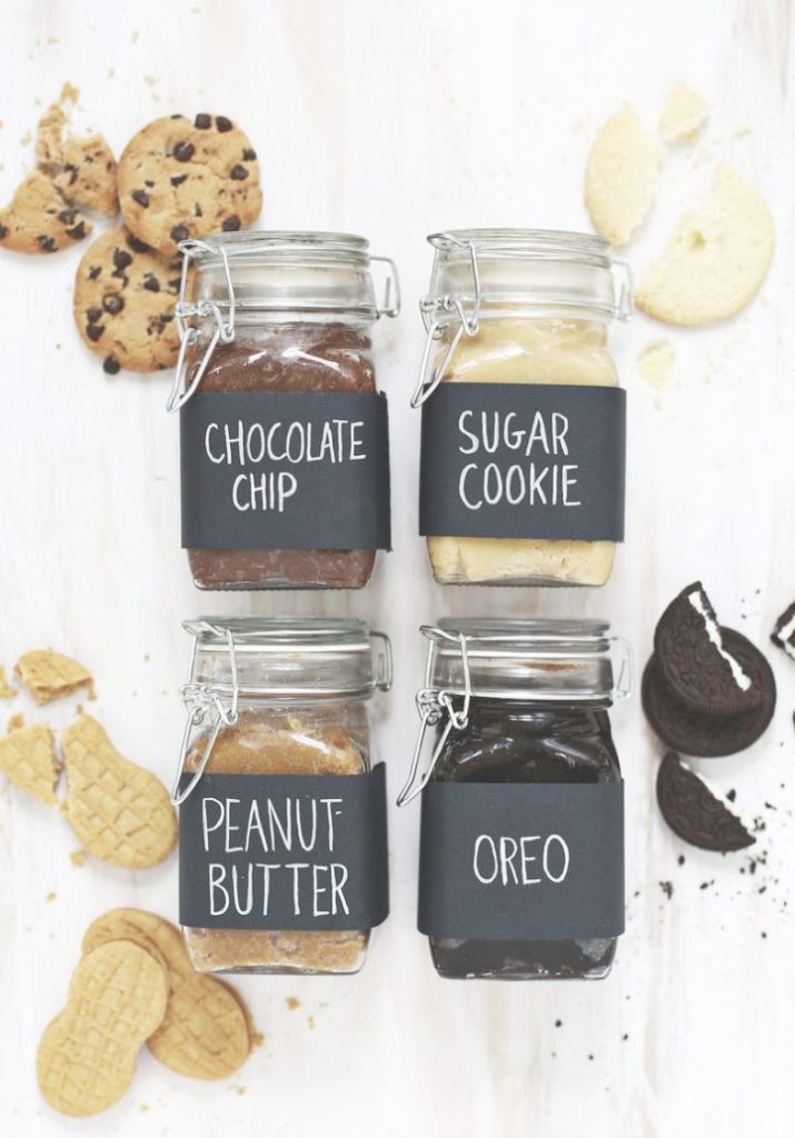 Four glass jars labeled Chocolate Chip, Sugar Cookie, Peanut Butter, and Oreo are filled with spreads, surrounded by various types of cookies on a light surface.