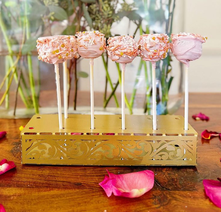 Six pink cake pops with sprinkles on sticks are displayed upright in a decorative gold holder on a wooden table with scattered rose petals.