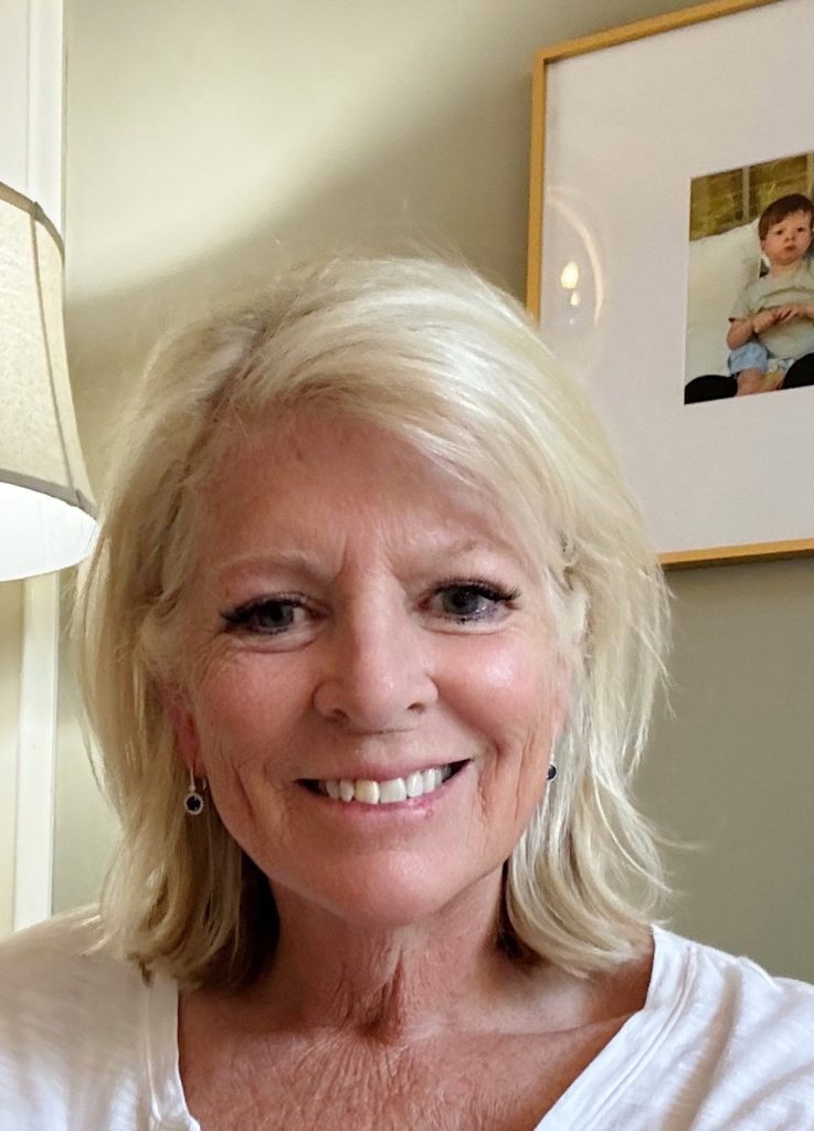 A woman with short, blonde hair and light skin smiles at the camera. She is indoors, wearing a white top, with a framed photo of a child visible on the wall behind her.