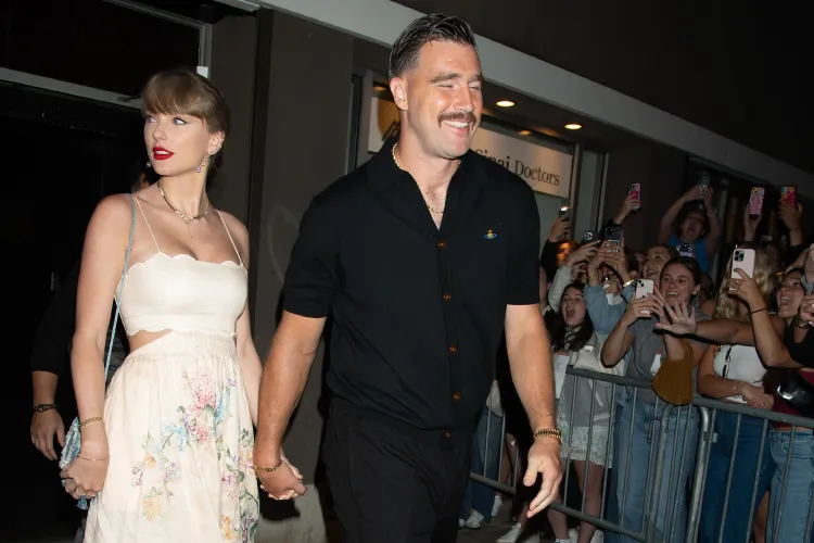 A woman in a white floral dress and a man in a black shirt hold hands while walking past a crowd of onlookers behind a barricade at night.