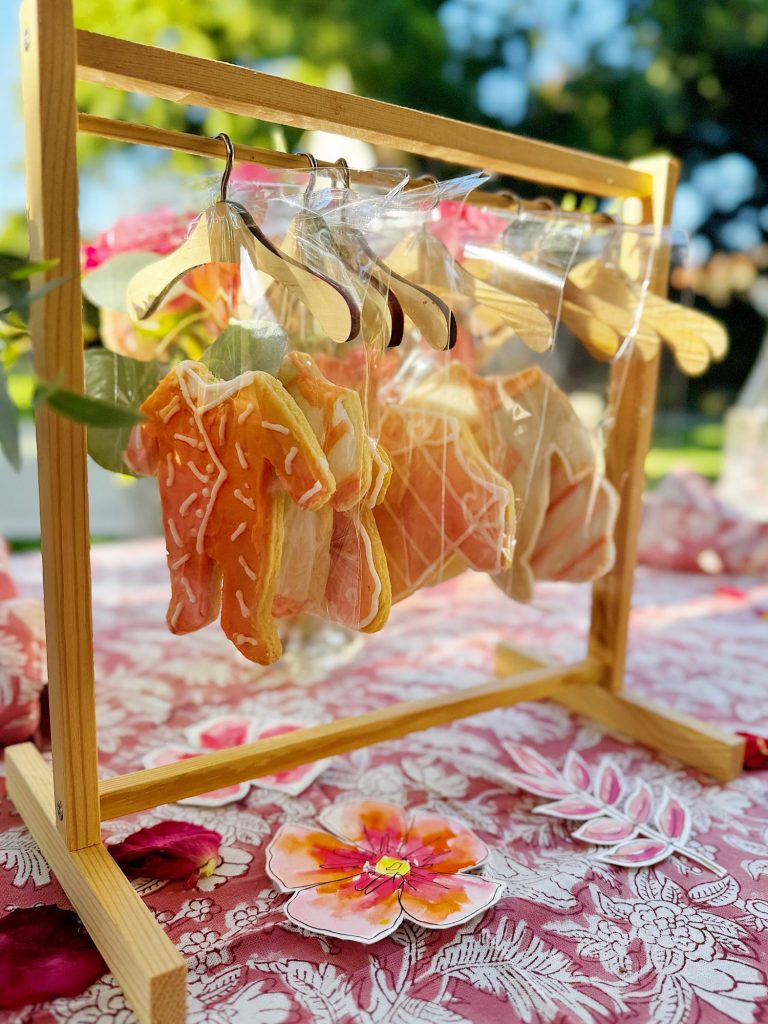 Decorated cookies shaped like clothing hang on miniature hangers under a wooden rack, displayed on a floral-patterned tablecloth outdoors.