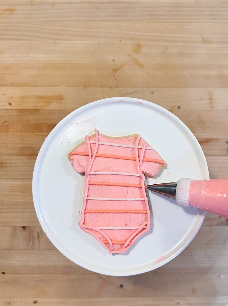 A sugar cookie shaped like a baby onesie is being decorated with pink icing on a white plate, using a piping bag.