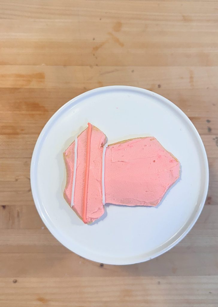 A cookie shaped like a pencil with pink icing, placed on a white plate atop a wooden surface.