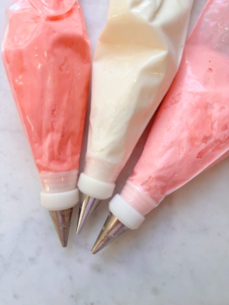 Three piping bags filled with pink, white, and light pink icing are arranged on a white surface, each with a metal decorating tip attached.
