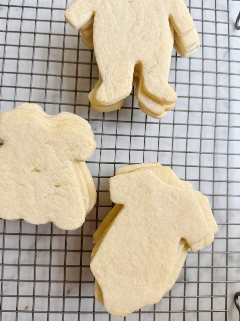 Three stacks of plain, uniced sugar cookies shaped like clothing items and a teddy bear rest on a metal cooling rack.