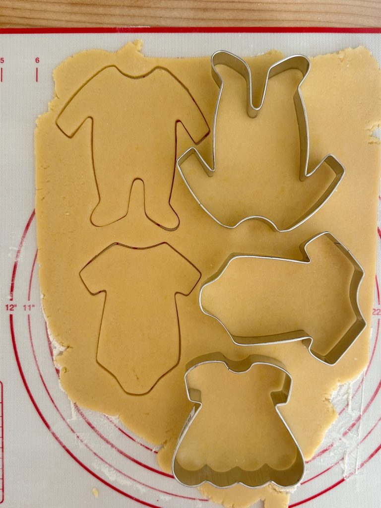 Cookie dough with four metal cookie cutters in the shapes of a onesie, a dress, a romper, and a bottle on a silicone mat.