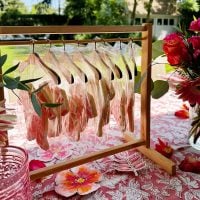 A wooden stand holds several bagged cookies on hangers atop a pink floral tablecloth, with flowers and glassware arranged nearby outdoors.
