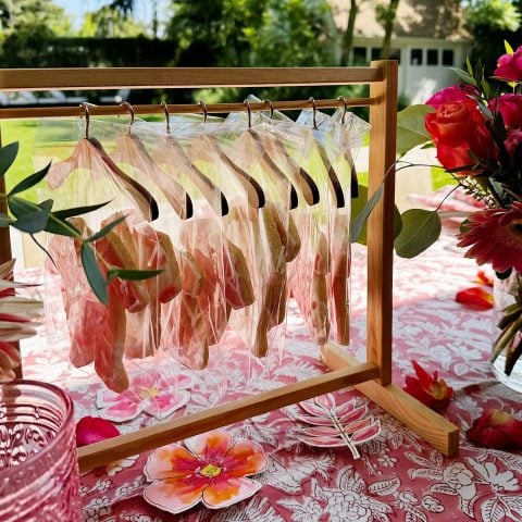 A wooden stand holds several bagged cookies on hangers atop a pink floral tablecloth, with flowers and glassware arranged nearby outdoors.