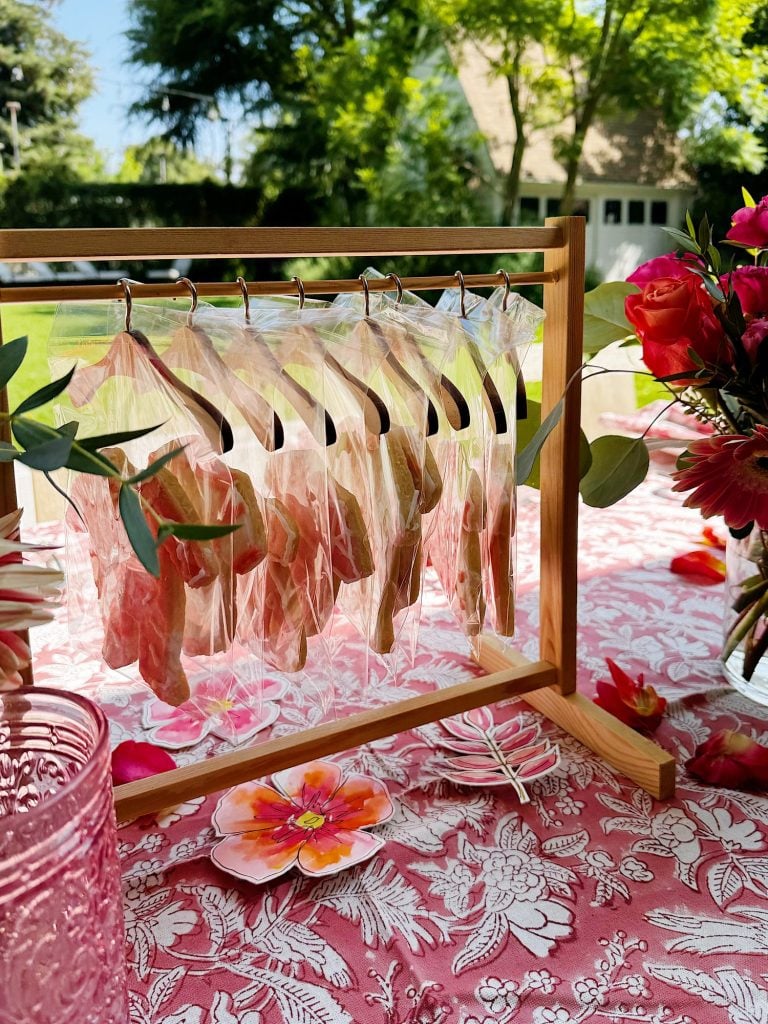 A wooden stand holds several bagged cookies on hangers atop a pink floral tablecloth, with flowers and glassware arranged nearby outdoors.
