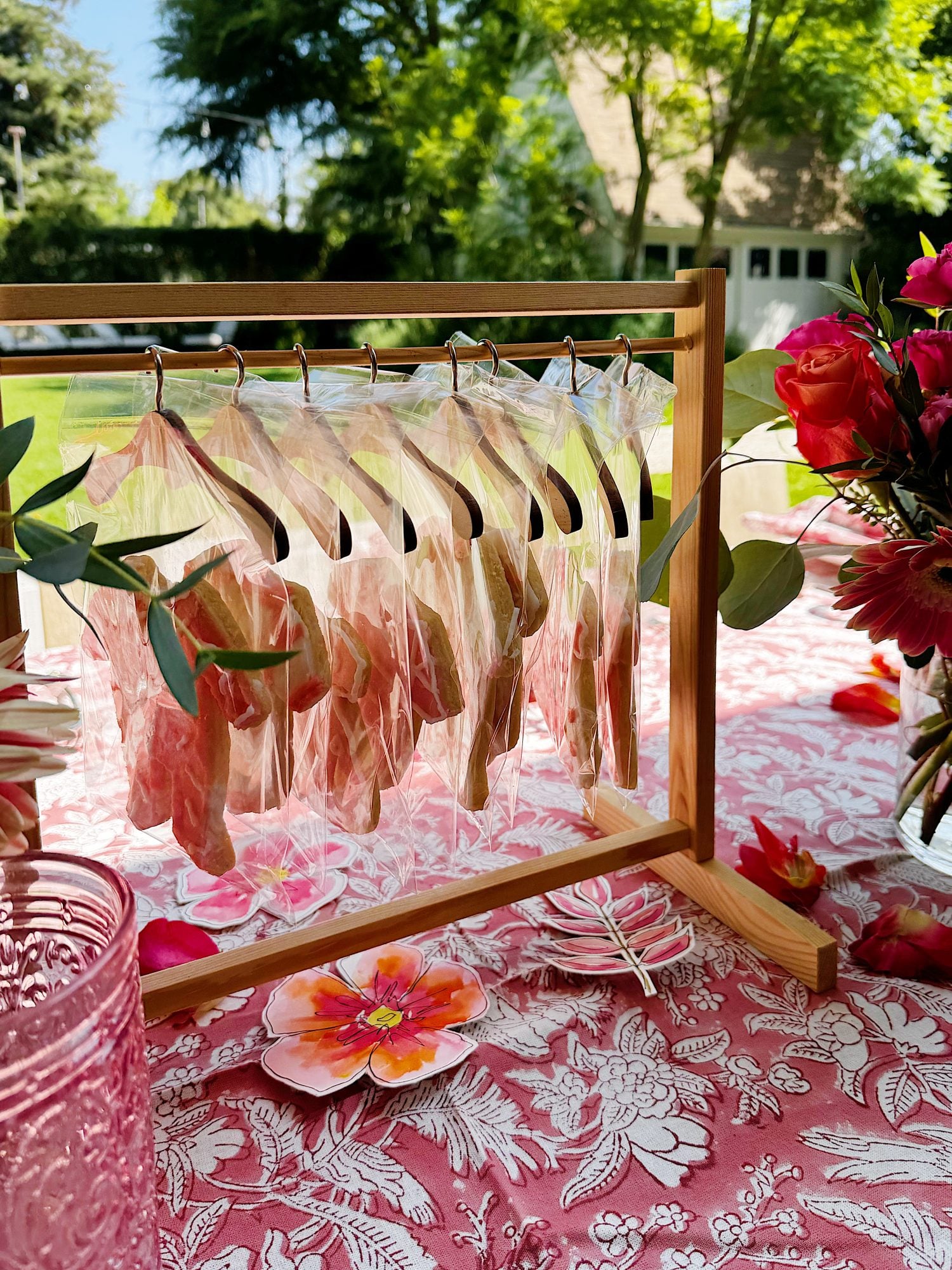 A wooden stand holds several bagged cookies on hangers atop a pink floral tablecloth, with flowers and glassware arranged nearby outdoors.