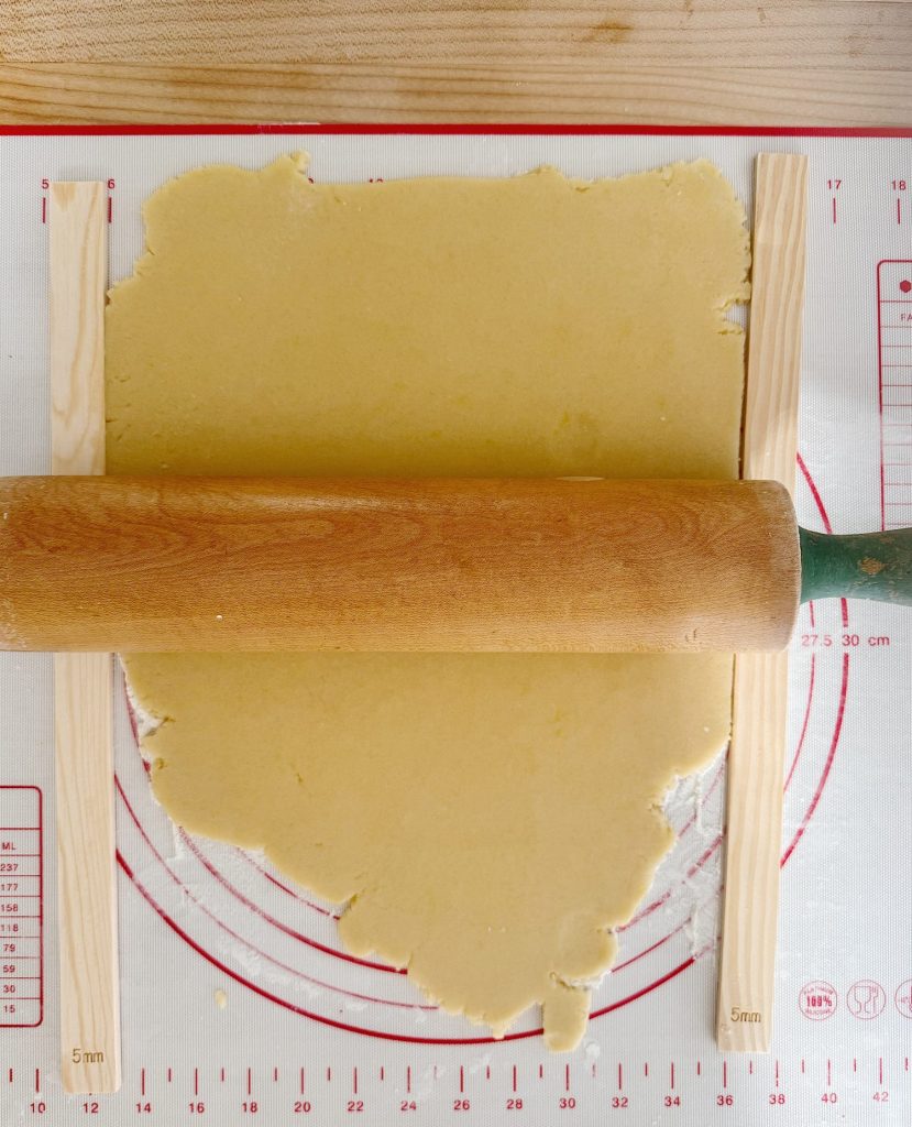 A rolling pin flattens dough between two wooden guides on a baking mat with measurement markings.