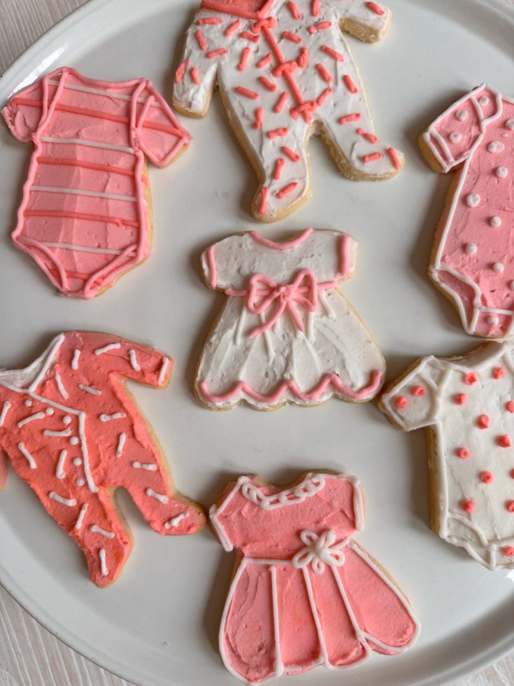 Plate of sugar cookies decorated with icing to resemble baby onesies and dresses in pink and white tones.