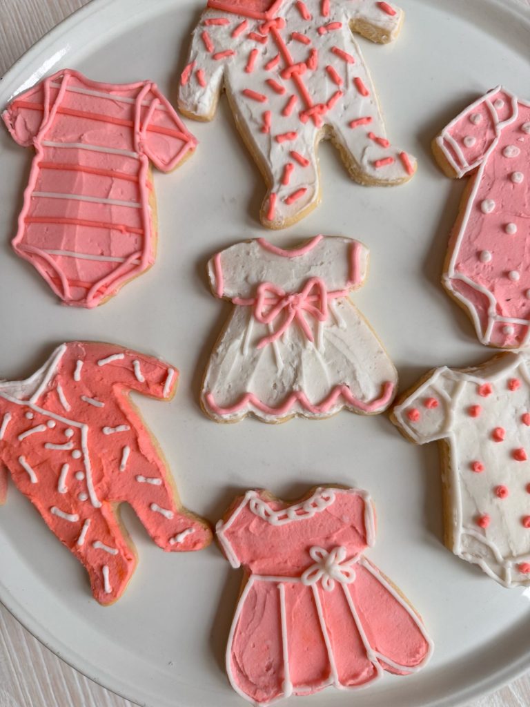 Plate of sugar cookies decorated with icing to resemble baby onesies and dresses in pink and white tones.